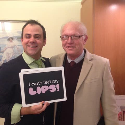 A smiling patient holding a humorous sign about dental anaesthesia alongside a practitioner